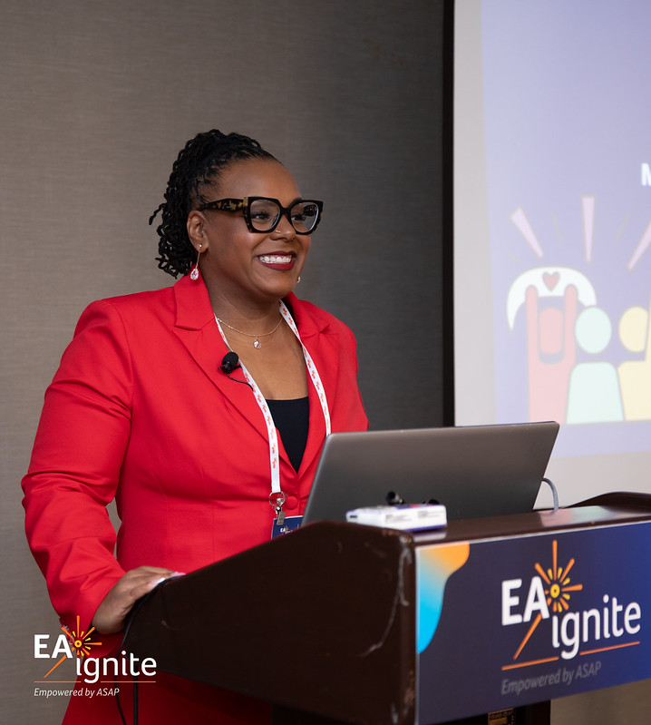 A woman in a red blazer stands at a podium labeled "EA Ignite," presenting in front of a screen with colorful graphics.