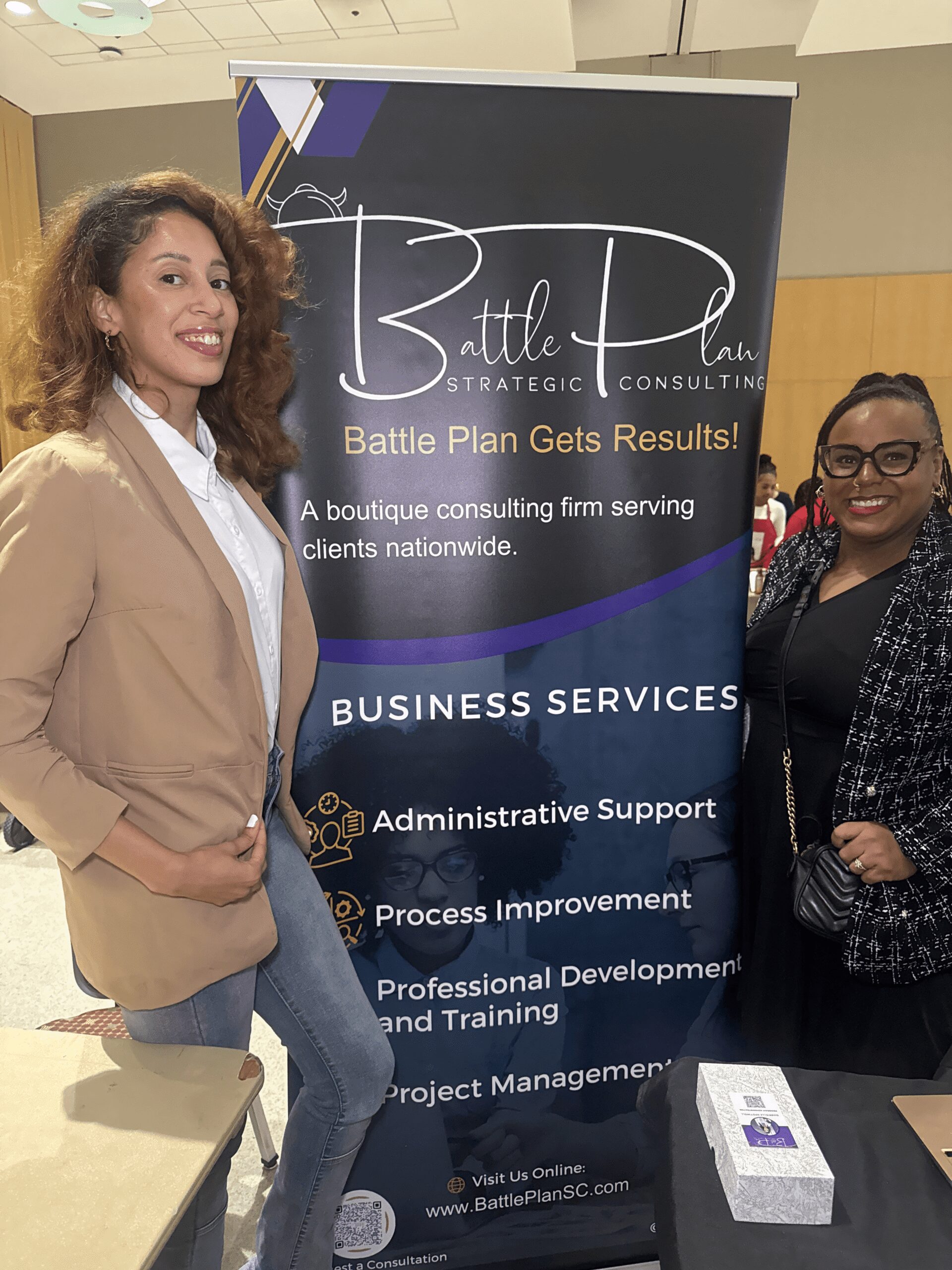 Two women stand beside a large vertical banner for Battle Plan Strategic Consulting, listing business services such as administrative support and process improvement.