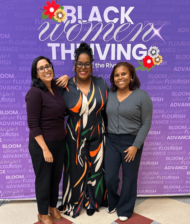 Three women stand smiling in front of a purple backdrop that reads "Black Women Thriving" with floral decorations on the sign.