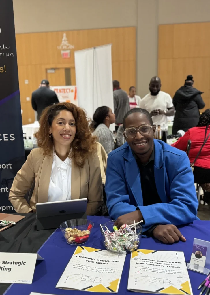 Two people sit at an event booth with informational flyers and a bowl of candy on the table in front of them; other attendees and booths are visible in the background.
